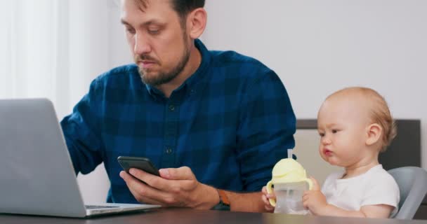 Mladý vousatý muž sedí doma u stolu s laptopem, píše na klávesnici notebooku, vzkazy na smartphonu Baby na vysoké dětské židli, nápoje z láhve a tlačí ji Muž otře stůl a kojenecké tváře — Stock video