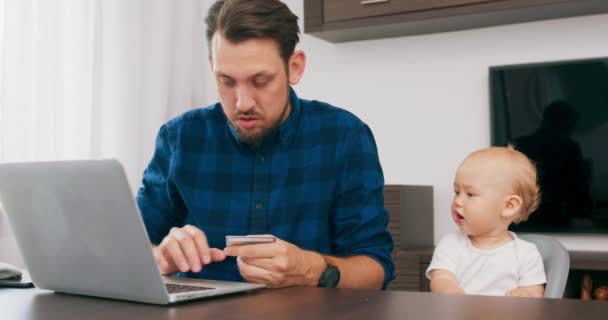 Homem barbudo jovem sentado na mesa em casa com laptop, segura um cartão de crédito na mão e escreve o número de cartões Bebê bonito senta-se na cadeira de bebê alta, e olhar para baixo com o pai câmera lenta — Vídeo de Stock