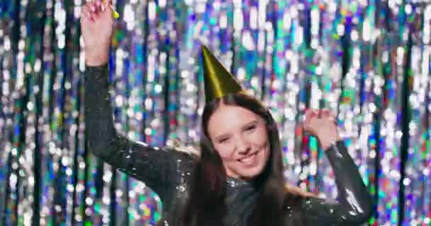 Ein junges hübsches Mädchen in einem Brokatkleid und einem Partyhut auf dem Kopf feiert ihren Geburtstag Sie tanzt und lacht Im Hintergrund hängt ein silbrig glänzender Vorhang — Stockvideo