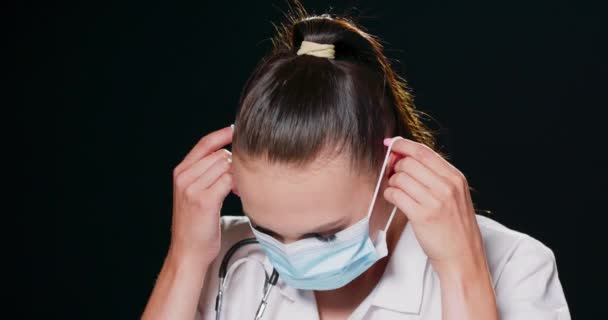 Primer plano de una profesional médica con una bata blanca de laboratorio con un estetoscopio alrededor del cuello poniéndose una mascarilla médica protectora contra un fondo negro de estudio con espacio para copiar — Vídeos de Stock