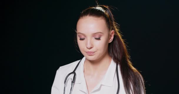 Portret młodej smutnej i zmęczonej lekarki w białym płaszczu z fonendoskopem Pracownica szpitala patrząca w kamerę i uśmiechnięta, studio, szare tło — Wideo stockowe