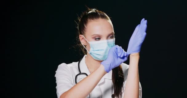 Vážná lékařka s obličejovou maskou, bílým laboratorním pláštěm a stetoskopem si nasadí chirurgické rukavice a podívá se na kameru na černém pozadí studia s kopírovacím prostorem — Stock video