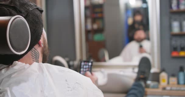 Голова молодого бородатого человека, который сидит на стуле парикмахера, лицом к зеркалу, с ногой на шкафу, держа смартфон Отражение в зеркале мужчины и женщины парикмахера, который держит — стоковое видео