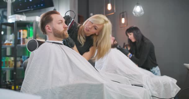 Homem barbudo jovem está sentado na cadeira barbeiro, com capa, de frente para o espelho, cortadores de barbeiro feminino seu cabelo com cortador de cabelo Atrás deles, outro barbeiro feminino, que se barba com barba de barbear de — Vídeo de Stock