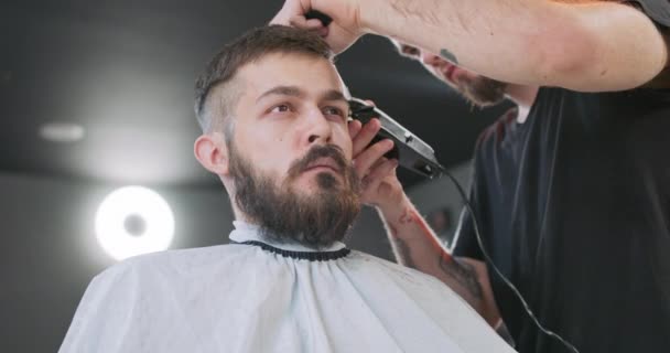 Pohled z dolní části obličeje mladého vousatého muže, který sedí na křesle holiče, čelem k zrcadlu, a samec vousatý holič střihač jeho vlasy přes ucho s střihačem vlasů a hřeben Šedý strop — Stock video