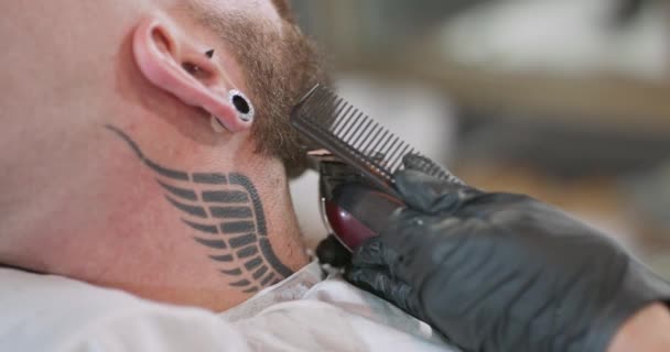 Vista por trás do pescoço e da cabeça do jovem barbudo, com brinco de túnel na orelha, e tatuagem de asa no pescoço, que está sentado na cadeira de barbeiro, e mãos em luvas de balck de barbeiro fêmea — Vídeo de Stock