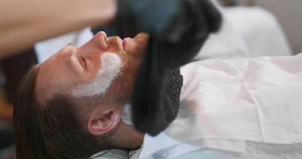 Zbliżenie ręce kobiety fryzjera, który owija czarny ręcznik na młodych brodatych mężczyzn twarz i szyję, z kremem do golenia — Wideo stockowe