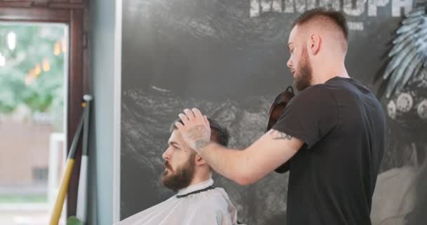 Homem barbeiro com espelho nas mãos, fica, atrás do cliente, mostra o resultado de seu trabalho ao jovem, que está sentado na cadeira barbeiro, com janela de capa, parede cinza no fundo — Vídeo de Stock