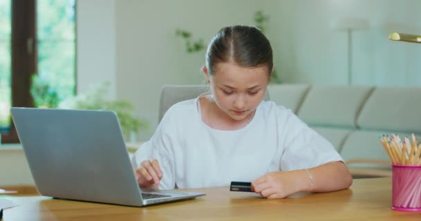 Linda chica adolescente en el escritorio, sosteniendo una tarjeta de crédito, escribe una tarjeta de datos en el ordenador portátil, haciendo e-shopping Notas, lápices, lámpara están en la mesa Plantas verdes, sofá, paredes blancas y una ventana en backgroung — Vídeos de Stock