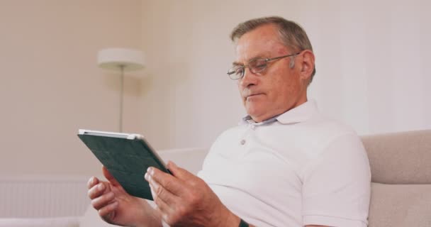 Un primer plano de un abuelo usando una tableta Hombre sentado en el sofá y leyendo nuevo en su tableta — Vídeo de stock