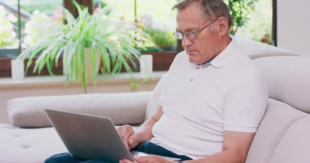 Старший пенсионер печатает на ноутбуке дома, на диване, в очках и работает с ноутбуком в гостиной. — стоковое видео