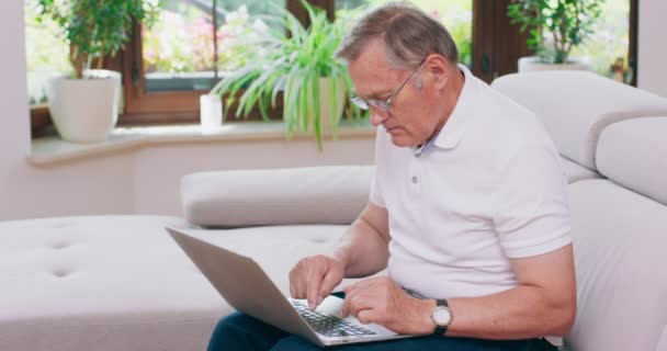 Egy nyugdíjas férfi egy laptopon dolgozik otthon egy kanapén. Idős nagyapa szemüveget visel és laptopon dolgozik a nappaliban. — Stock videók