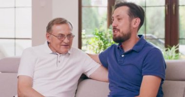 Mutlu iki nesil erkek aile, yaşlı yaşlı yaşlı baba ve gülümseyen yetişkin oğul sohbetin tadını çıkarıyorlar sohbetin tadını çıkarıyorlar arkadaşça olumlu sohbetin tadını çıkarıyorlar evdeki kanepede oturuyorlar.