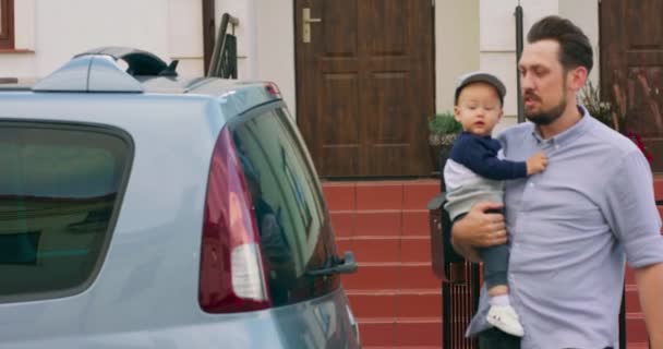 Mladý vousatý otec nese chlapečka a dává ho do dětské autosedačky v rodinném autě, zatímco jde ven ze vstupních schodů svého domu Zpomalený pohyb — Stock video