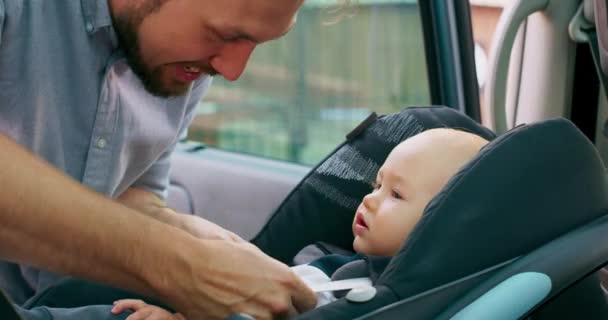 車の中のカメラクローズアップ赤ちゃんの男の子は車の中の赤ちゃんの車の座席に座っています若いひげを生やした父親は赤ちゃんがシートベルトを固定チェックし、彼に話しますスローモーション — ストック動画