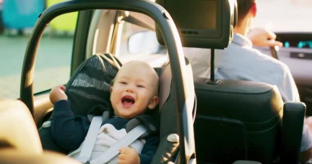 Κοντινό πλάνο γέλιο αγοράκι κάθεται στο κάθισμα του αμαξιού μωρό στο πίσω κάθισμα στο εσωτερικό του αυτοκινήτου, ενώ το αυτοκίνητο είναι ιππασία κατά μήκος του δρόμου Πατέρας πίσω του οδηγεί αργή κίνηση — Αρχείο Βίντεο