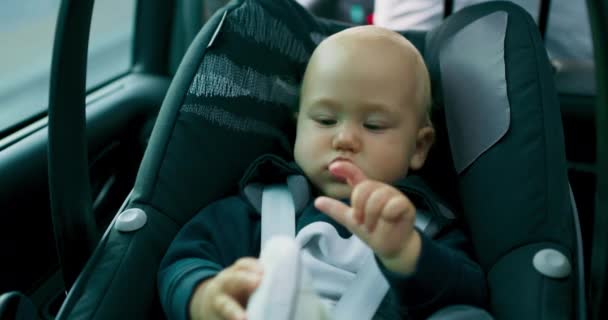 Closeup face and hands of baby boy in the baby car seat on the rear seat of car while car is riding along the road Baby pulls up left foot with shoe then looks at fingers of his hands Slow motion — Stock Video
