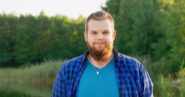 Портрет улыбающегося молодого человека с рыжей бородой на открытом воздухе в лесу возле озера — стоковое видео