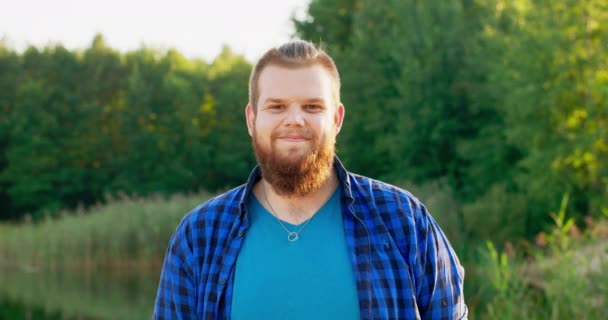 Retrato de um jovem sorridente com uma barba vermelha ao ar livre em uma floresta perto de um lago — Vídeo de Stock