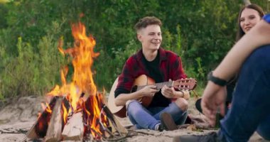Genç çocuğun arkadaşlarıyla şakalaşıp nehir kenarında piknik yaptığı gün batımına yakından bakın. Genç kız gitar çalacak.