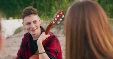 Nehir kenarında piknik yaparken arkadaşlarıyla konuşan genç bir çocuğun görüntüsü. Gün batımında boş vakit geçiriyor. Genç kız gitar çalacak.