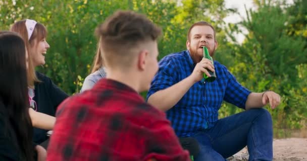 Portret młodego brodatego chłopca z nadwagą żartującego ze swoimi przyjaciółmi i pijącego piwo podczas pikniku nad brzegiem rzeki Jasny zachód słońca spędzającego wolny czas — Wideo stockowe