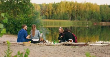 Bir grup arkadaş gün batımında kumsalda şenlik ateşi etrafında parti veriyorlar. Sakallı hippi adam şakalar yapıyor. Bayram günbatımının sıcacık renkleri.