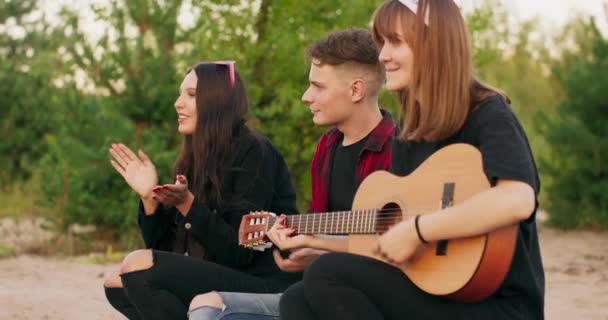 Młodzi mężczyźni i kobiety śpiewają piosenki do gitary odpoczywając przy ognisku i ciesząc się muzyką i dobrym towarzystwem w letnie noce Drzewa są widoczne — Wideo stockowe
