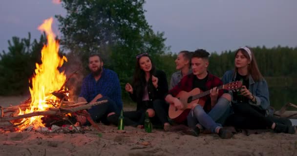 Pique-nique de jeunes avec feu de joie sur la plage le soir Des amis joyeux chantent des chansons et jouent de la guitare Slowmotion shot — Video