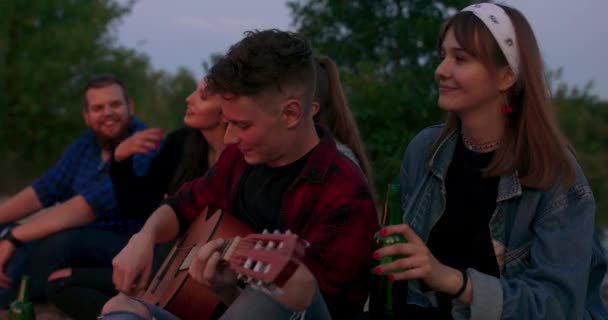 Hipster homme jouant de la guitare acoustique et chantant chanson avec des amis voyageurs à grand feu de joie au camp de nuit dans la forêt Groupe de personnes se refroidissant au feu le soir camping près du lac — Video