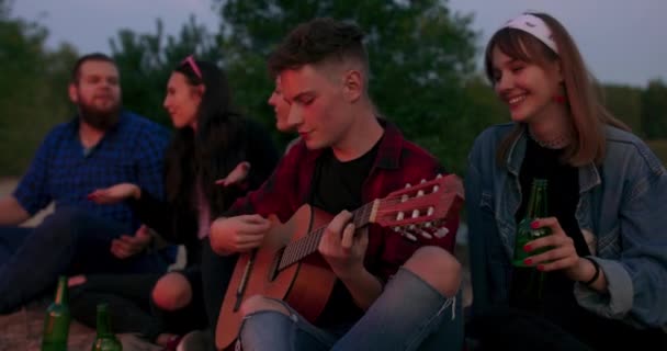 Unga män och kvinnor sjunger sånger till gitarren som vilar runt lägerelden och njuter av musik och gott sällskap på sommarnatten Träd och sjö syns — Stockvideo