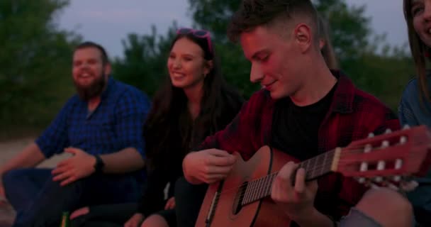 Filmisk vy genom ljus gyllene låga på ung tonåring man spelar gitarr nära brasa ler vänner vid lägereld — Stockvideo