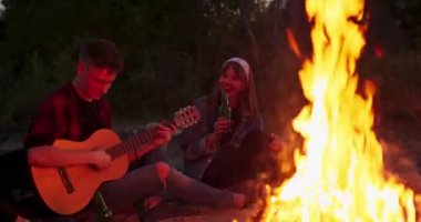 Genç bir çocuk arkadaşlarıyla kamp ateşi etrafında otururken gitar çalar.