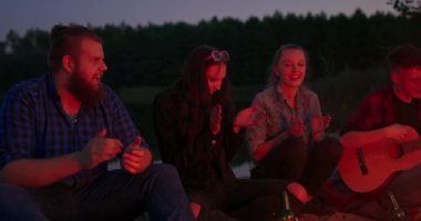 Yakışıklı genç çocuk gitar çalarken arkadaşları komik şarkılar söylüyor ve gülüyor ve el çırpıyor. Müzik doğa ve eğlence kavramı.