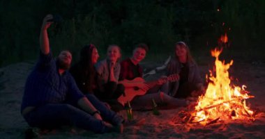 Genç erkekler ve kadınlar kamp ateşi etrafında dinlenerek gitarla şarkılar söylüyor ve yaz gecelerinde müzik ve güzel arkadaşlıkların tadını çıkarıyorlar. Ağaçlar ve göl görülebiliyor.