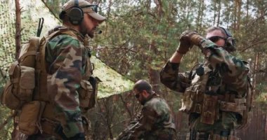 Askeri üniformalı bir grup asker, geçici bir orman üssünde harekete geçmeye hazırlanıyor. Woodland askeri üniformalı, sakallı, orta yaşlı bir asker, haberleşme ekipmanlarını onun üzerine giyiyor.