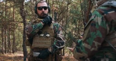 Woodland askeri üniformalı ve şapkalı sakallı orta yaşlı bir asker kafasına mikrofon takıp komutanla iletişimi kontrol ediyor.
