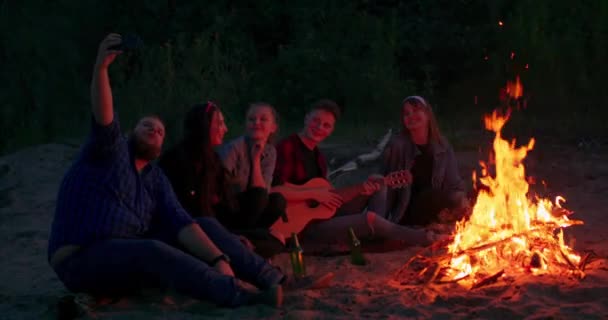 Fiatal férfiak és nők dalokat énekelnek a gitárnak a tábortűz körül pihen, és élvezik a zenét és a jó társaságot a nyári éjszakán Fák és tó látható — Stock videók