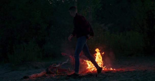 Mans átugrik tábortüzet a strandon éjszaka Fiatal turista nő ül a gitár körül tűz az erdőben este — Stock videók