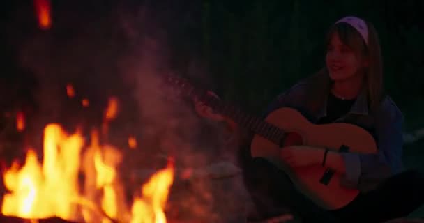 Vista cinematográfica a través de la llama dorada brillante en la joven adolescente tocando la guitarra y cantando cerca de la hoguera Amigos sonrientes en la hoguera — Vídeos de Stock