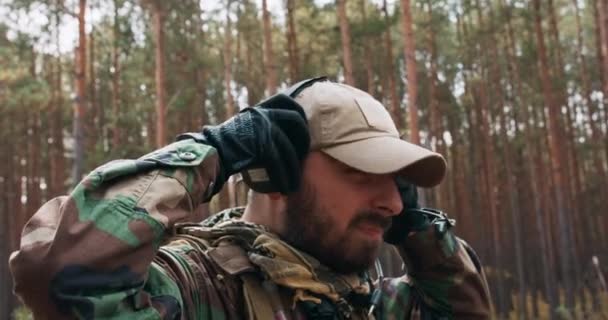 Un soldado barbudo de mediana edad con un uniforme militar de Woodland y gorra se pone auriculares tácticos con un micrófono sobre su cabeza y comprueba la comunicación con el comandante. — Vídeos de Stock