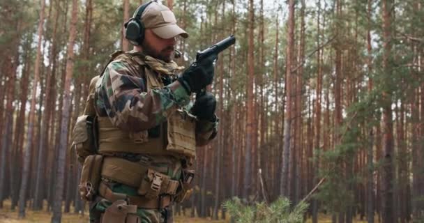 Ein bärtiger Soldat mittleren Alters in Woodland-Uniform und Mütze mit taktischen Kopfhörern und Mikrofon auf dem Kopf überprüft seine Waffe, bevor er zur Tat schreitet. — Stockvideo