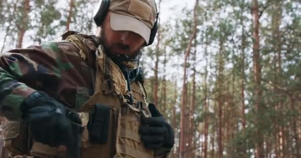 Um soldado barbudo de meia-idade de uniforme militar Woodland e boné em fones de ouvido táticos com um microfone na cabeça guarda uma arma — Vídeo de Stock