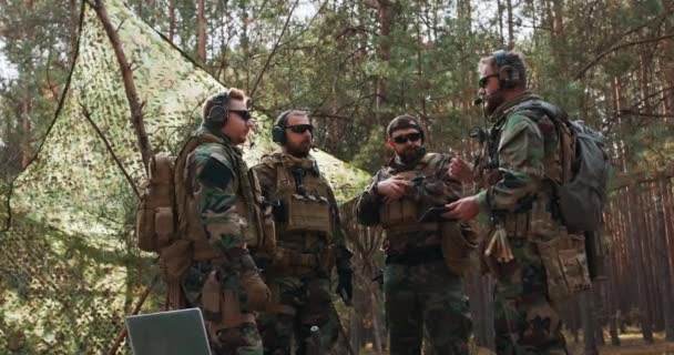 Grupa brodatych żołnierzy w średnim wieku w mundurach i kamizelkach taktycznych omawia plan działania i przygotowuje się do akcji w tymczasowej bazie leśnej. — Wideo stockowe