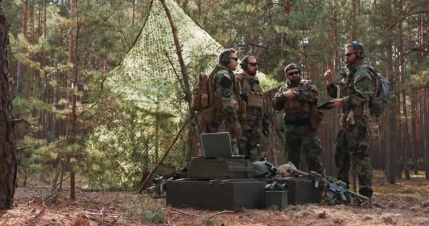 Um grupo de soldados barbudos de meia-idade em uniformes e coletes táticos discute o plano de ação e se prepara para a ação em uma base florestal temporária. — Vídeo de Stock