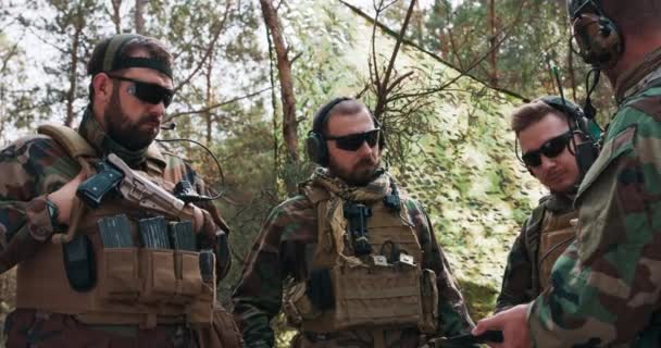 군복 과 전술 조끼를 입은 중년의 군인들 이 군사 용 태블릿을 사용하는 사령관 이 임시 숲 기지에서 행동 계획에 대해 논의하고 준비하고 있다. — 비디오