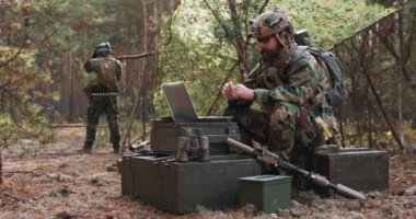 Taktik yelek giymiş, askeri üniformalı sakallı bir komutan kafasında bir miğferle, geçici bir orman üssünün bilgisayarında sohbet ederken arka planda üssü koruyan bir askeri görebilirsiniz.