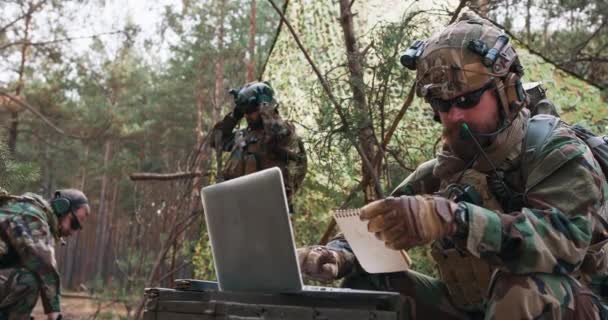 Brodaty dowódca brygady w mundurze wojskowym z hełmem na głowie sprawdza współrzędne z oficjalnego notesu i analizuje je na laptopie wojskowym. — Wideo stockowe