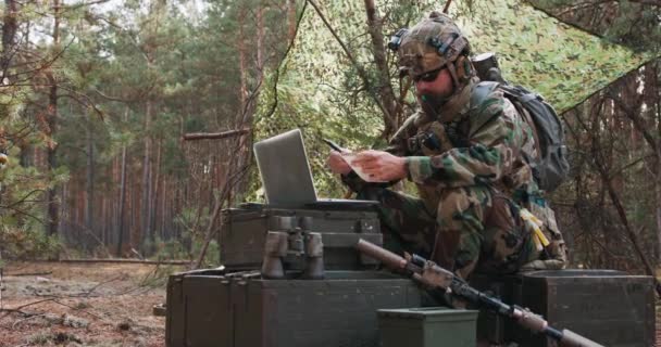 Een bebaarde brigade commandant in een militair uniform met een helm op zijn hoofd controleert de coördinaten vastgelegd in zijn officiële notitieboekje en analyseert ze op een militaire laptop in een tijdelijke bosbasis — Stockvideo