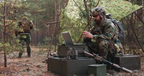 Un commandant barbu en uniforme militaire dans un gilet tactique avec un casque sur la tête bavardant en ligne sur un ordinateur portable à une base forestière temporaire En arrière-plan, vous pouvez voir un soldat gardant la base — Video